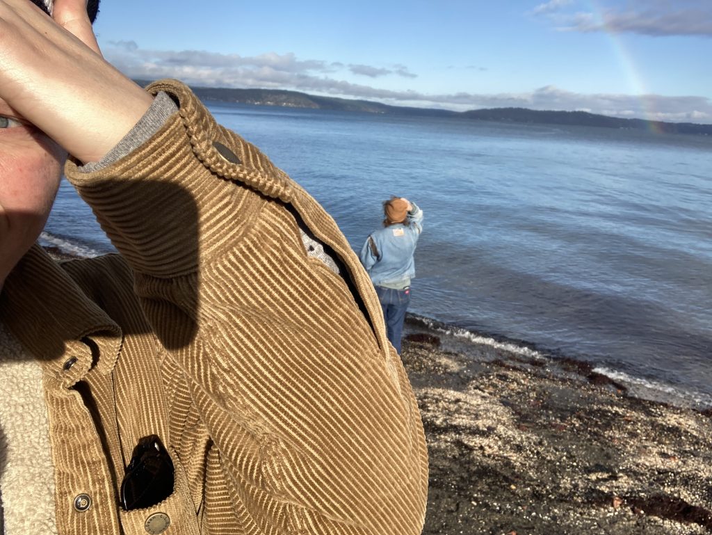 a selfie showing part of my face. my arm, in a corduroy jacket, is raised to block the sun. in the background a person looks out over water. there are mountains in the distance and a rainbow rising from the water into the sky.