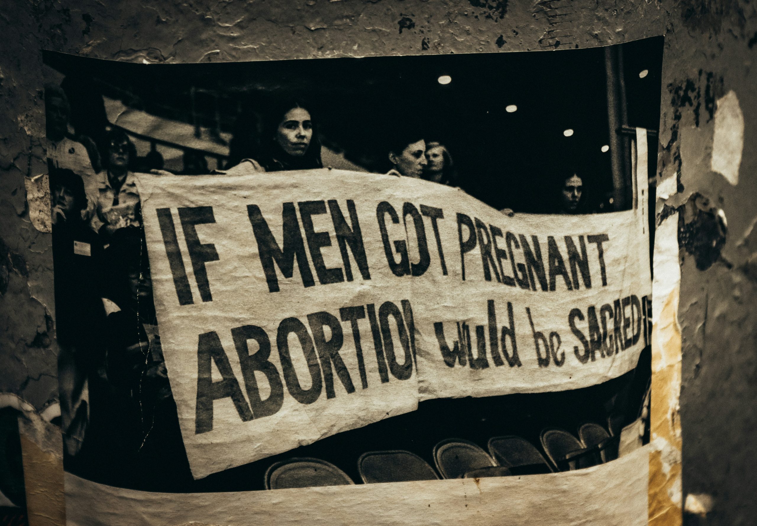 A photograph of a black-and-white photograph: in the photograph a row of women standing holding a large white banner that says "IF MEN GOT PREGNANT, ABORTION would be SACRED."